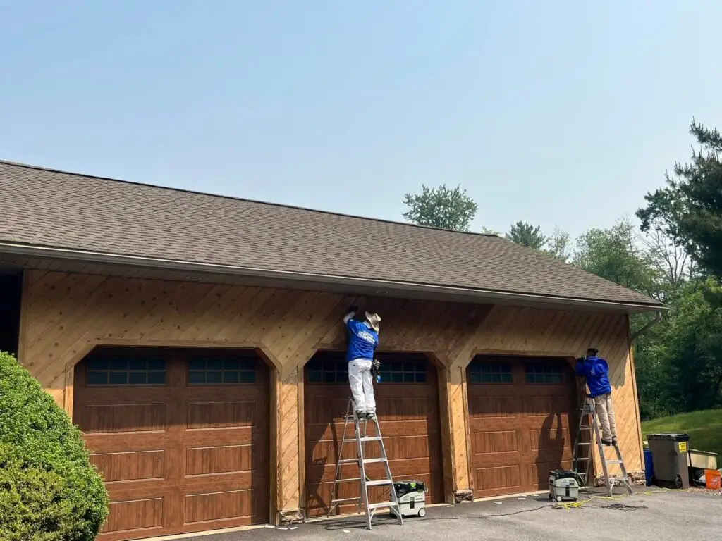 Two painters from Davis Painting on ladders applying finish to a large wooden garage with three doors under a clear blue sky.