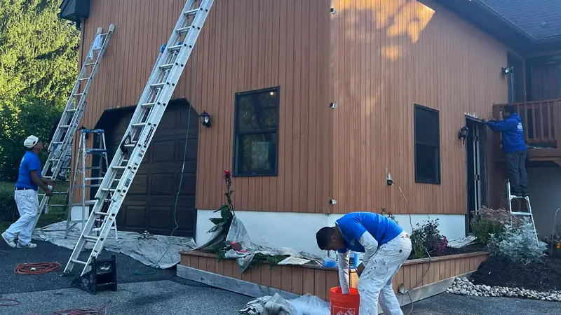 Three workers from Davis Painting repair siding on a two-story house with brown walls, using ladders and various tools.