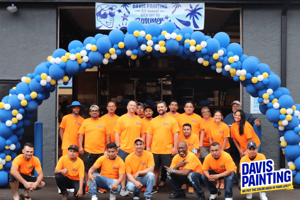 A group of about 20 people, wearing matching orange shirts, pose for a group photo under a blue and white balloon arch outside a building. A banner overhead reads "Davis Painting" with a summer-themed design. The "Davis Painting" logo is visible in the bottom right corner.