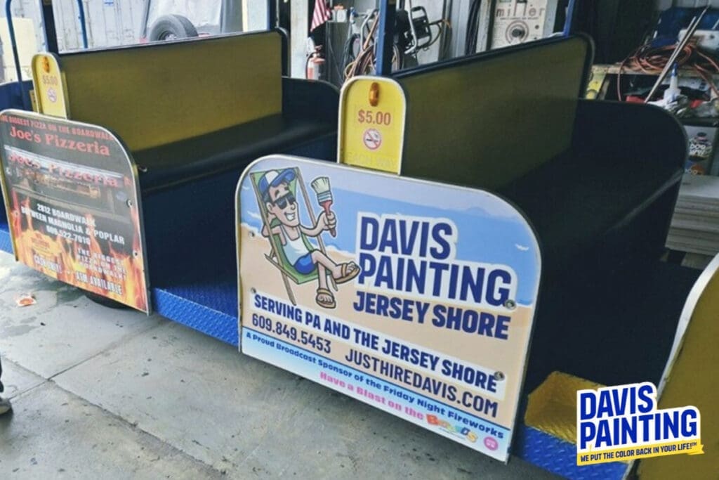 Two side-by-side miniature train rides are parked in an indoor space. The train on the right advertises "Davis Painting: Jersey Shore" with contact information, a cartoon figure painting with a brush, and links to their social media. A sign indicates the ride costs $5.00.