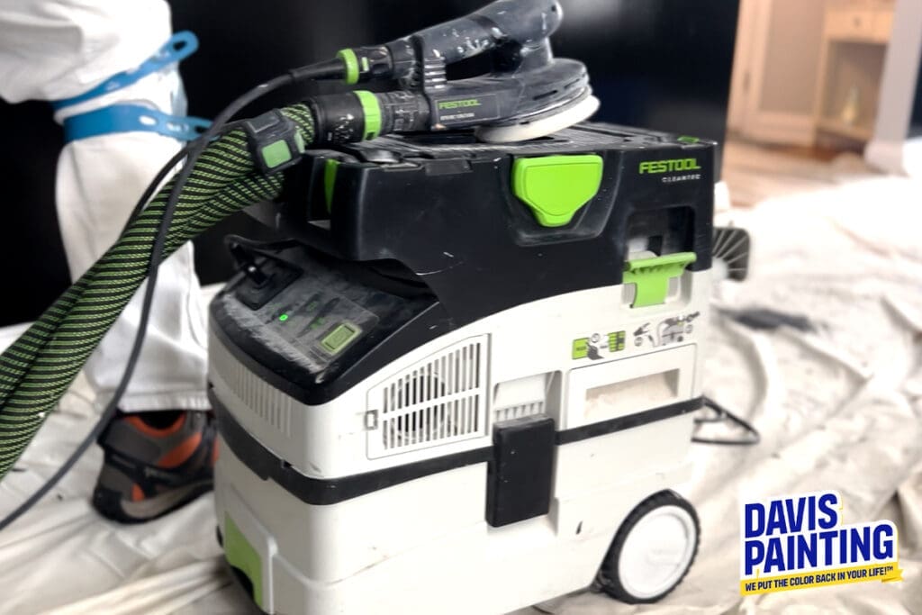 A Festool dust extractor is connected to a sander. A worker in white overalls and a blue safety harness is partially visible behind the equipment. The machine is on a white drop cloth in a room with light-colored furniture. The Davis Painting logo is in the bottom right corner.