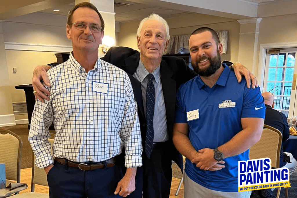 Three men stand together indoors, smiling at the camera. Each man wears a name tag on his shirt—one is in a checkered shirt, another in a suit and tie, and the third in a blue polo shirt with "Davis Painting" printed on it. A "Davis Painting" sign is seen at the bottom right.