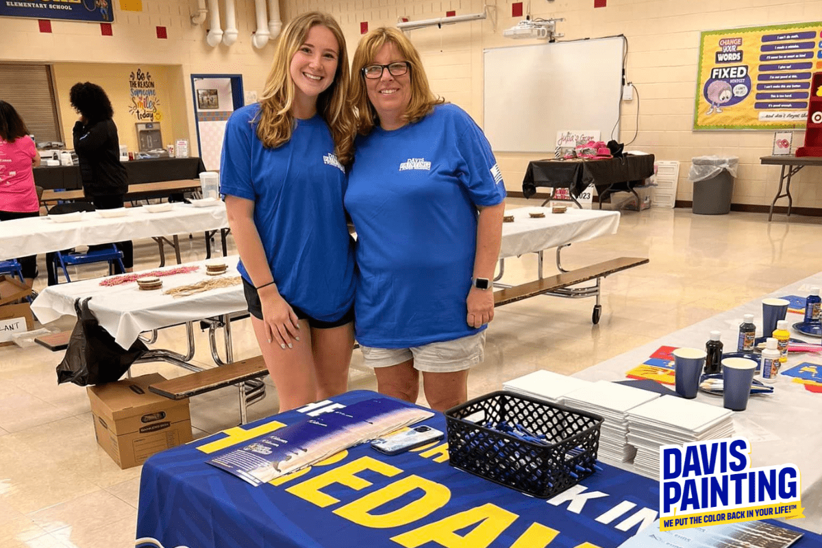Two women, both wearing blue "FREEDAY" shirts, smile for a photo in a bright room with tables and supplies in the background. One woman has long hair and is standing, the other has short hair and glasses. A "Davis Painting" sign is displayed on the table in front of them.