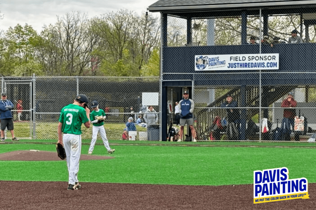 A youth baseball game is in progress on a field. A player in white pants and a green jersey with number 3 is at the pitcher's mound. Spectators and other players are in the background. A scoreboard with "DAVIS PAINTING" sponsorship is visible on the right.