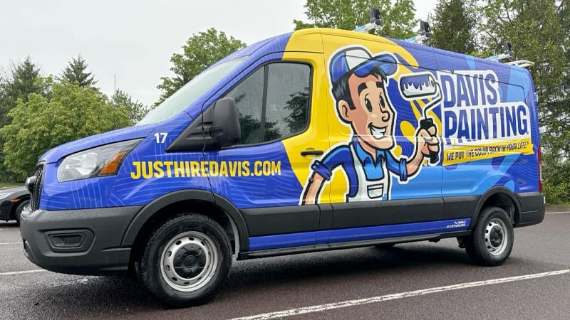 A blue and yellow van for "Davis Painting" featuring a cartoon painter holding a paint roller. The text "JUSTHIREDAVIS.COM" and "WE PAINT THE WORLD A BETTER PLACE!" is prominent on the sides, inviting new careers in painting. The van is parked in a lot with trees in the background.