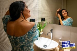 A woman stands in a bathroom, styling her hair while looking in the mirror. She is wearing a blue, floral-patterned dress. The sink counter holds a glass vase with green leaves and white flowers. The Davis Painting logo and slogan appear in the bottom right corner.