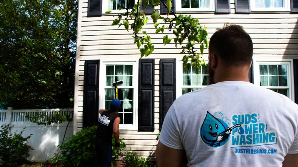 Two people are seen cleaning the exterior windows of a house. One person is wiping a window with a squeegee, while the other, wearing a "Suds Power Washing" shirt, observes. Inside, there's prep work for interior painting visible through the windows. The house is white with black shutters and greenery and a white fence in the background.