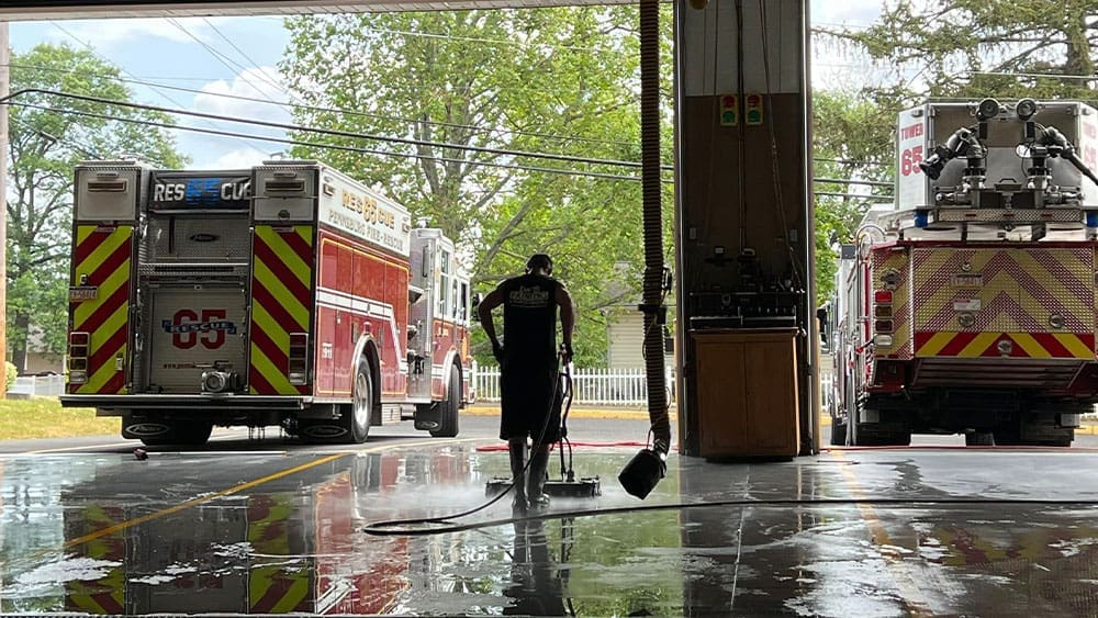 A firefighter is cleaning the floor inside a fire station, using a hose to spray water. Two fire trucks with emergency lights and equipment are parked inside, alongside an area designated for interior painting projects. The station’s bay doors are open, revealing a sunny day with trees and a white fence outside.