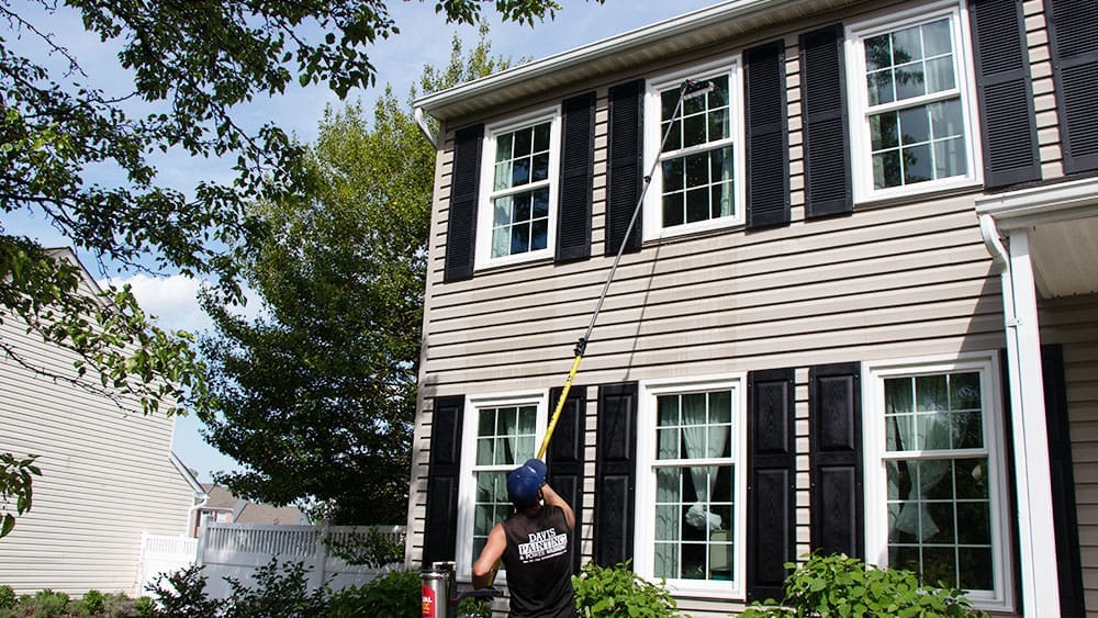 A person wearing a black tank top and cap is using a long telescopic pole to clean the second-floor windows of a beige house with black shutters. The house, which has just undergone interior painting, is surrounded by green trees, and the sky is partly cloudy.