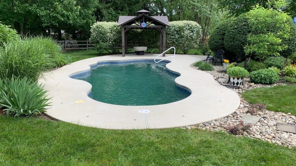 A serene backyard pool scene with a freeform in-ground swimming pool surrounded by a neatly trimmed lawn. A small pavilion with a bench and a decorative hanging, reminiscent of an exquisite interior painting, is located at the far end, and the area is framed by lush greenery and bushes.