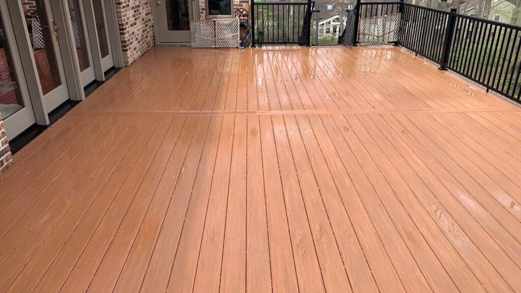 A spacious wooden deck glistening with rainwater is bordered by black metal railings and attached to a brick house with glass doors, inviting you inside where expert interior painting enhances the warm, cozy ambiance. In the background, trees and additional houses are visible through the railings.