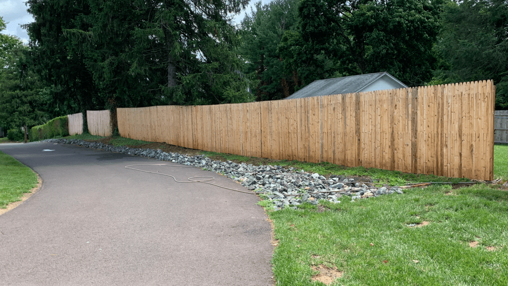 A paved pathway runs alongside a tall wooden fence, bordered by a strip of rocks. A coil of hose lies on the ground near the fence. Trees and greenery are visible in the background, enclosing the residential area, creating an inviting ambiance perfect for contemplating future interior painting projects.