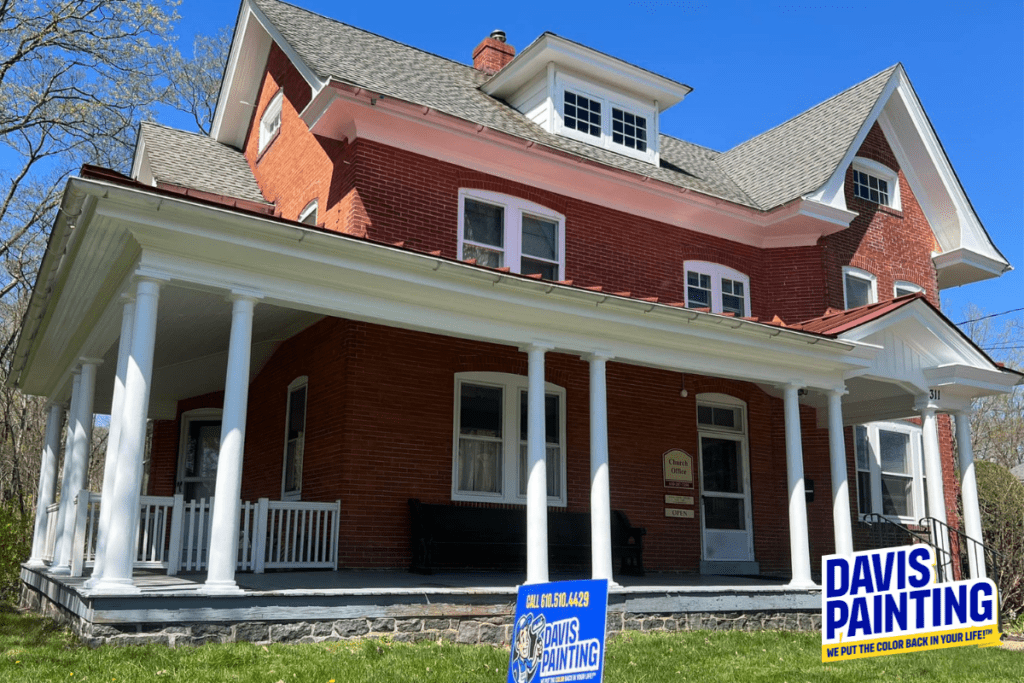 A large red brick house with a white porch, several windows, and a signboard by the door. Two signs on the lawn read "DAVIS PAINTING" with a slogan underneath, showcasing their expertise in residential painting. The sky is clear and blue.