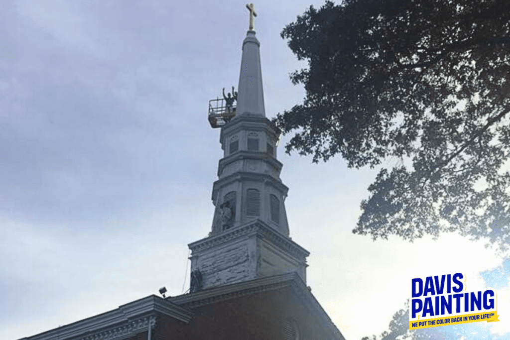 A tall church steeple under a cloudy sky, with a person on a lift performing maintenance near the top. The Davis Painting logo and tagline "We Put the Color Back in Your Life!" are visible in the bottom right corner, showcasing their expertise in both residential painting and larger projects.