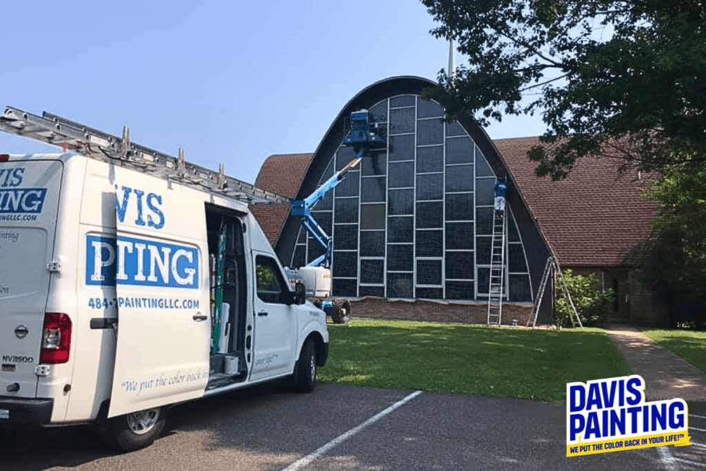 A white van with "Davis Painting LLC" branding is parked in front of a building with a large, curved roof and expansive windows. A worker on a blue lift is painting the building—a prime example of their expert residential painting services. The company's sign and slogan are displayed prominently in the bottom right corner.