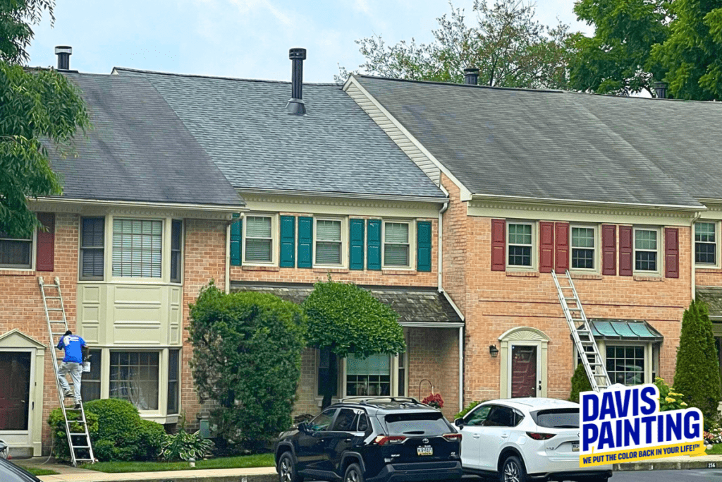 A worker in blue overalls paints the shutter of a window on a townhouse using a ladder on the left, while another ladder leans against a townhouse on the right. The houses are brick with colorful shutters. A Davis Painting sign, reminiscent of their renowned church painting projects, is visible in the bottom right corner.