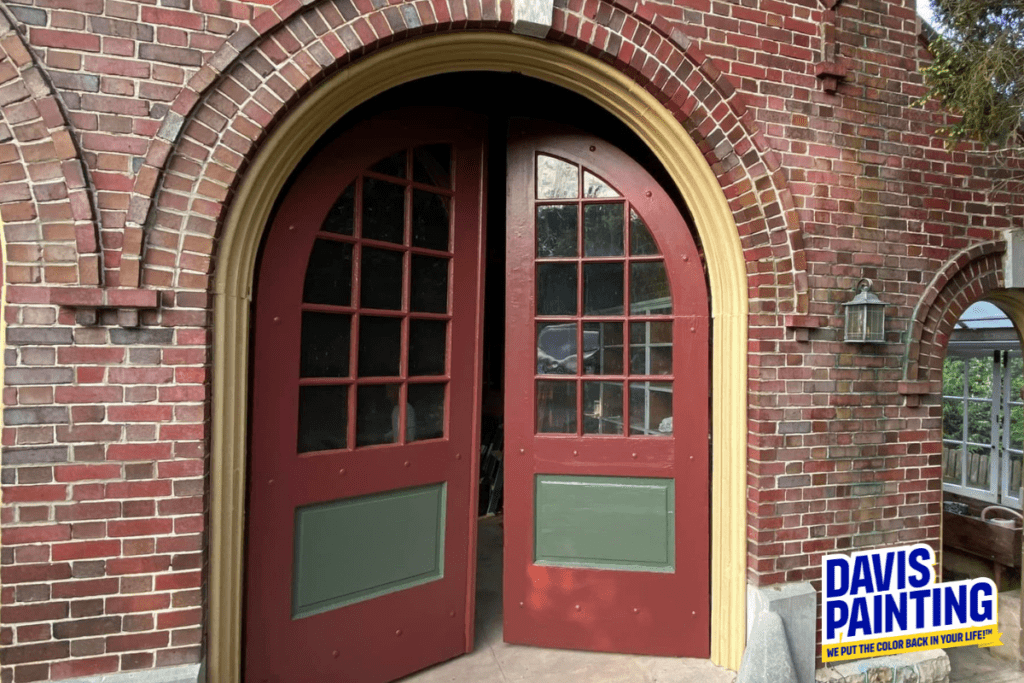 A pair of red arched doors with green panels are set within a brick building, resembling the entrance to a quaint church. The doors are partially open, revealing a glimpse into the interior. Beside the doors, a wrought iron lantern is mounted on the wall. A "Davis Painting" sign is visible in the lower right corner.