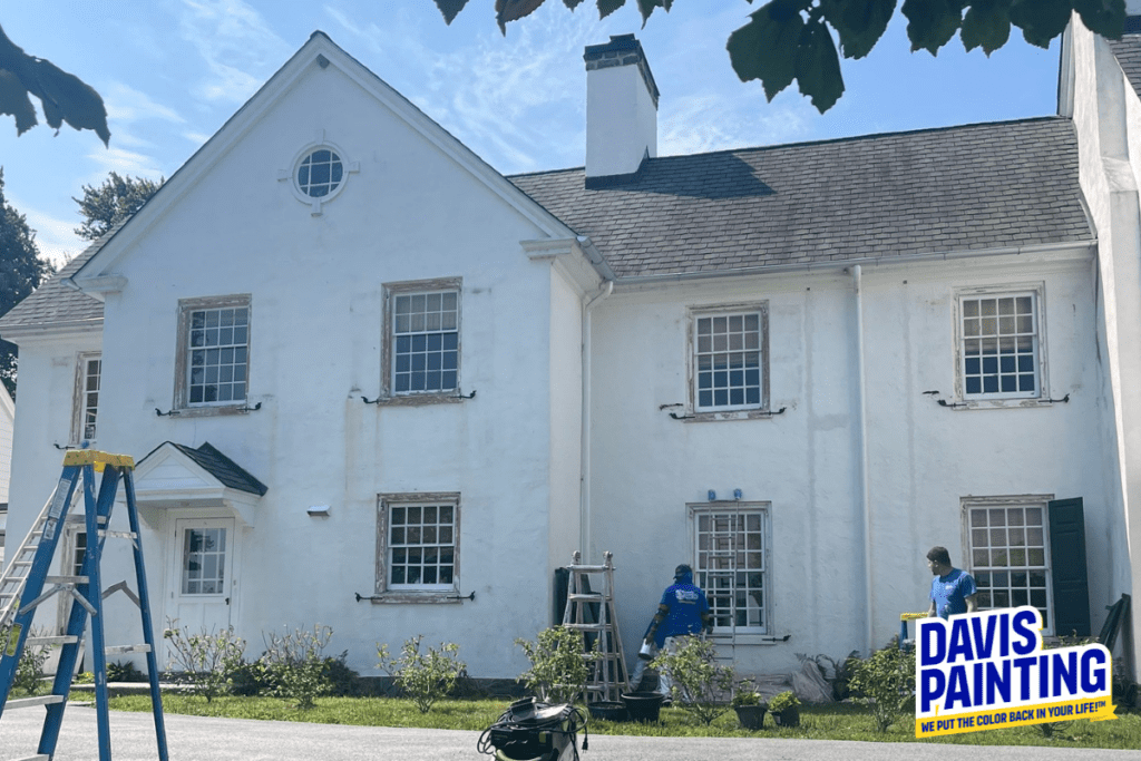 A white, two-story house is being painted by two workers using ladders to reach higher areas. Various painting supplies are on the ground. A sign in the bottom right corner reads "Davis Painting: We put the color back in your life!"—experts also trusted in church painting.