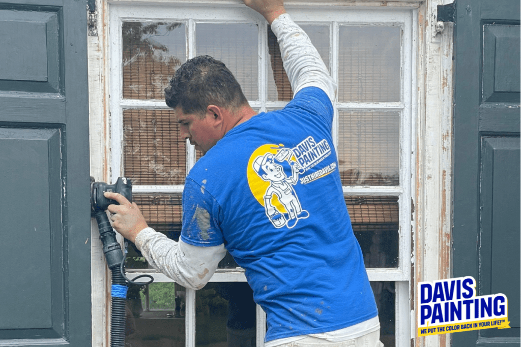 A man wearing a blue "Davis Painting" t-shirt is sanding a window frame with a power tool. The white frame, featuring multiple panes, stands out. His shirt and a graphic on the bottom right display the logo and slogan, "We Put the Color Back in Your Life!" showcasing their expertise, whether it's home or church painting.