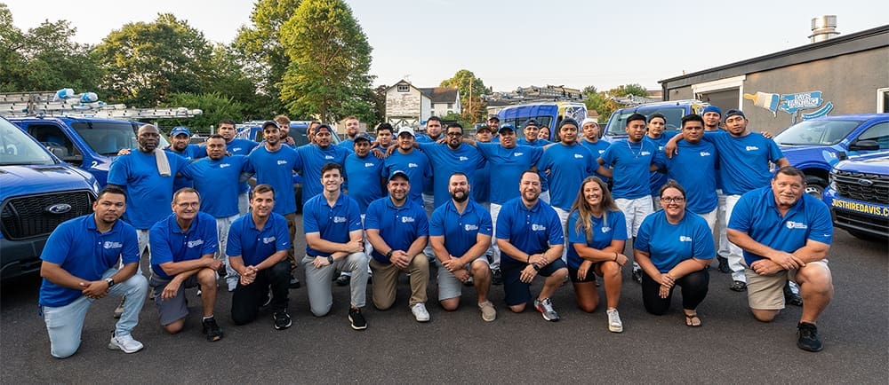 A large group of people wearing matching blue shirts and posing for a photo outdoors, perhaps for a company gallery. Some are standing while others kneel in front. Several blue trucks and greenery are visible in the background, emphasizing their unity as a team or company staff.