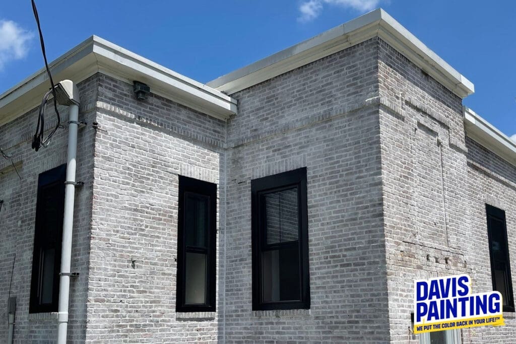 A two-story building with freshly painted light-gray brick exterior and black-framed windows. The flat roof contrasts with the clean, updated look of the walls. In the lower right corner, a logo reads "DAVIS PAINTING: WE PUT THE COLOR BACK IN YOUR LIFE!"—specializing in church painting.