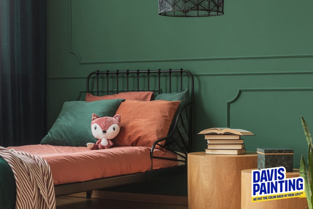 A cozy bedroom with green walls features a black metal bed frame, coral and dark green pillows, and a fox plush toy. Next to the bed is a round wooden nightstand holding books and a plant. With an eye for historical restoration, "Davis Painting" has its logo at the bottom right of the image.