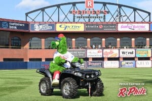 A green mascot in a baseball uniform rides an ATV on a baseball field, with a stadium wall and numerous advertising signs in the background. The mascot is wearing a red and blue cap, and the scene takes place on a sunny day. Text reads "Proud Partner of R-Phils." It’s as vibrant as a Davis painting restores churches.