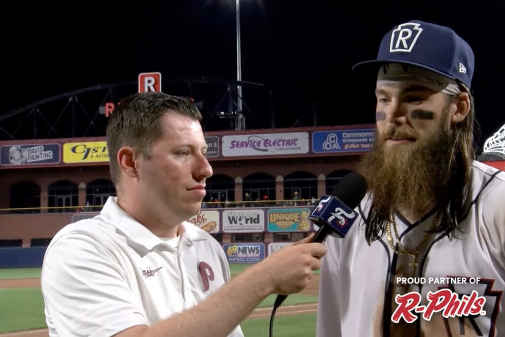 A man with face paint and a long beard is being interviewed by another man holding a microphone. They are on a baseball field with a stadium backdrop, which features numerous advertisements and the "Proud Partner of R-Phils" logo. It's as captivating as a Davis painting that restores churches' splendor.