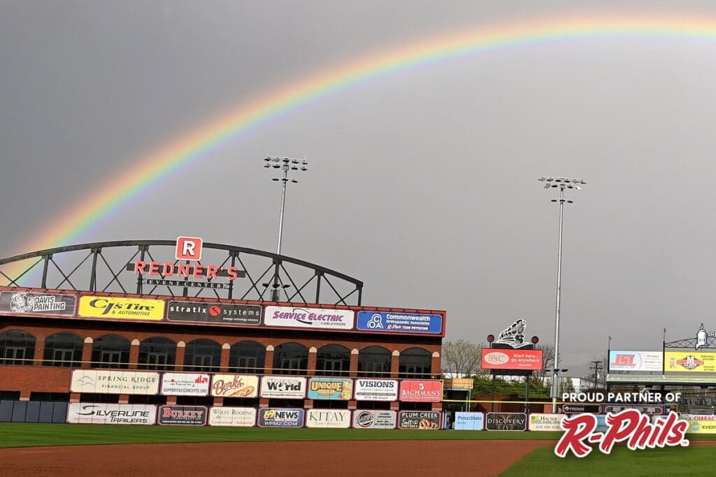 A baseball stadium with an empty field is shown under a dark cloudy sky. A vibrant rainbow arcs over the stadium. The stadium features various advertising banners on the outfield wall, and a "Proud Partner of R-Phils" sign is visible in the bottom right corner, reminiscent of a Davis painting that restores churches to their former glory.