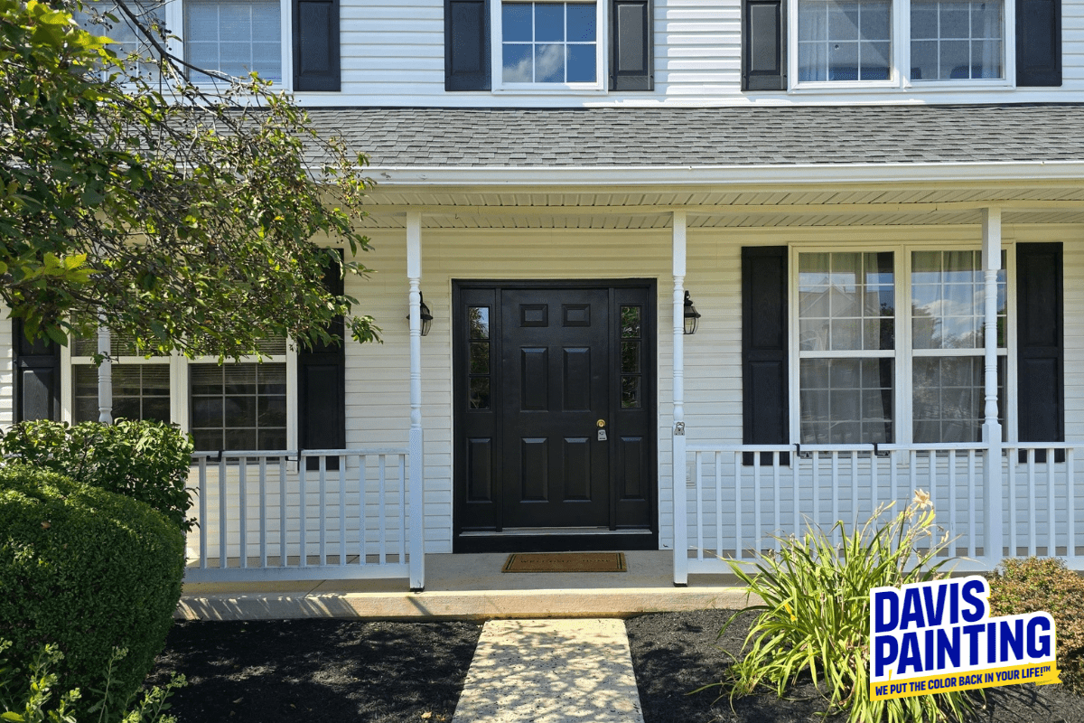 A two-story house with white siding and black shutters. The front door has a dark, polished finish and is flanked by two windows. A “Davis Painting” sign is visible in the bottom right corner near neatly trimmed bushes and plants adjacent to a concrete pathway, all done without the most common painting mistakes.