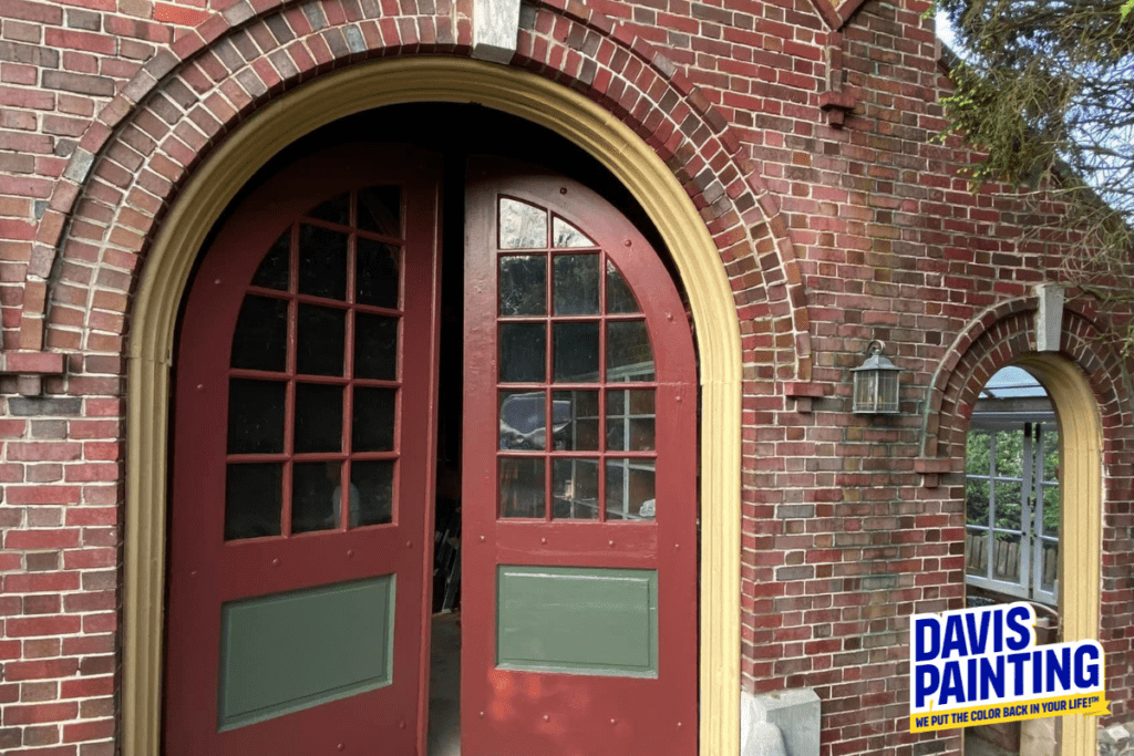 A red double door with window panes and green panels, set in a brick wall, is partly open. The entrance features an archway and glass panes. A sign on the bottom right reads, "Davis Painting," with the tagline "We put the color back in your life!"—just remember to wait between coats of paint.