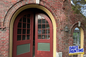 A red double door with window panes and green panels, set in a brick wall, is partly open. The entrance features an archway and glass panes. A sign on the bottom right reads, "Davis Painting," with the tagline "We put the color back in your life!"—just remember to wait between coats of paint.