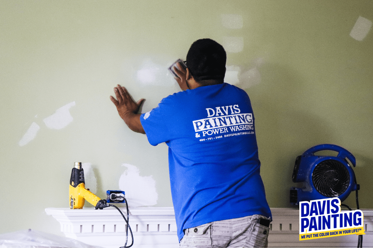 A person in a blue "Davis Painting" shirt sands a green wall with white patches in a room, preparing it for the next layer of oil vs. water-based paint. A heat gun and an industrial fan are placed on a white mantel nearby. The "Davis Painting" logo is visible in the bottom right corner.