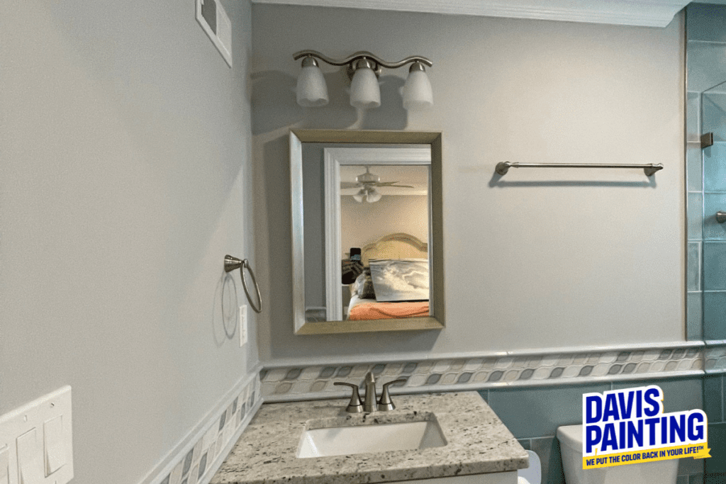 Bathroom interior with a granite countertop and rectangular mirror above the sink. A three-light fixture is mounted above the mirror. A towel bar is on the right wall. The Davis Painting logo is in the lower right corner.