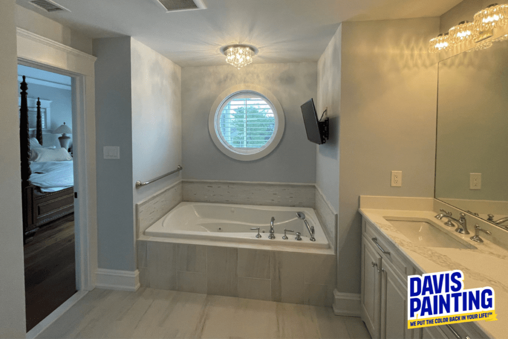 A modern bathroom with a jacuzzi tub under a round window, a TV mounted on the wall, and a sink with a large mirror. A logo in the corner reads "Davis Painting: We Put the Color Back in Your Life!.