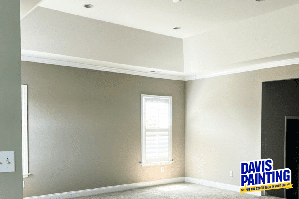 A freshly painted room with beige walls and white crown molding. Sunlight streams through a window with white shutters. The carpet is light gray. A logo in the corner reads, "Davis Painting: We put the color back in your life.