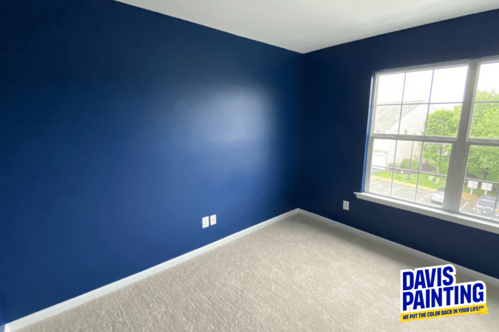 An empty room with dark blue walls and a light beige carpet. A window on the right side lets in natural light, showing a view of a neighborhood. The logo "Davis Painting" is in the bottom right corner.