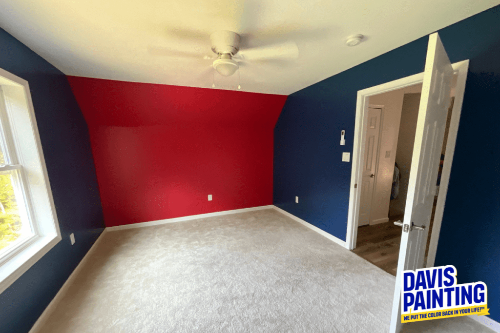 A newly painted room with vibrant red and blue walls, a white ceiling fan, and light beige carpet. Sunlight streams in through a window, and the door is open to an adjacent area. A "Davis Painting" logo is in the bottom right corner.