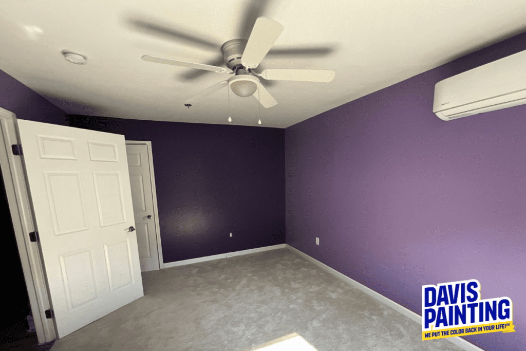 A room with purple walls and light gray carpet, featuring a white ceiling fan and an air conditioning unit. The logo "Davis Painting: We Put the Color Back in Your Life" is in the bottom right corner. An open door is visible.