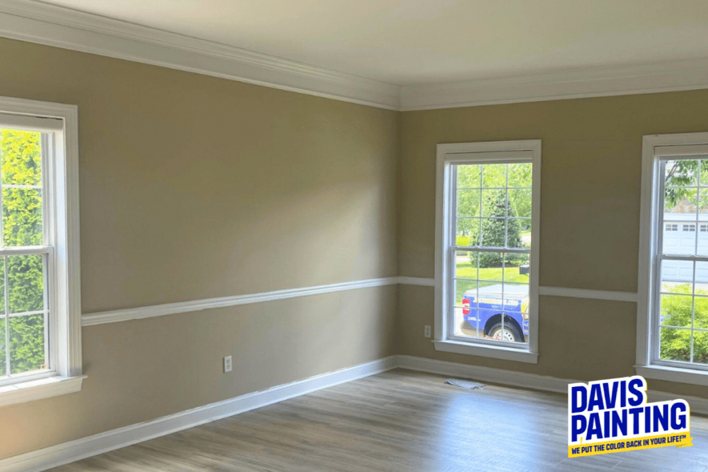 A brightly lit, freshly painted room with beige walls and white trim, featuring two large windows showing a sunny day outside. The floor is light-colored hardwood. A "Davis Painting" logo is in the bottom right corner.