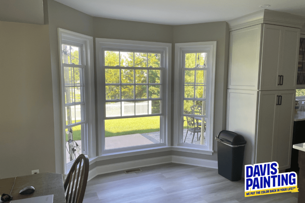 A brightly lit room featuring a large bay window with a view of a sunny garden. The room has light gray walls, a wooden table with chairs, a trash bin, and a logo in the corner for Davis Painting, showing their slogan.