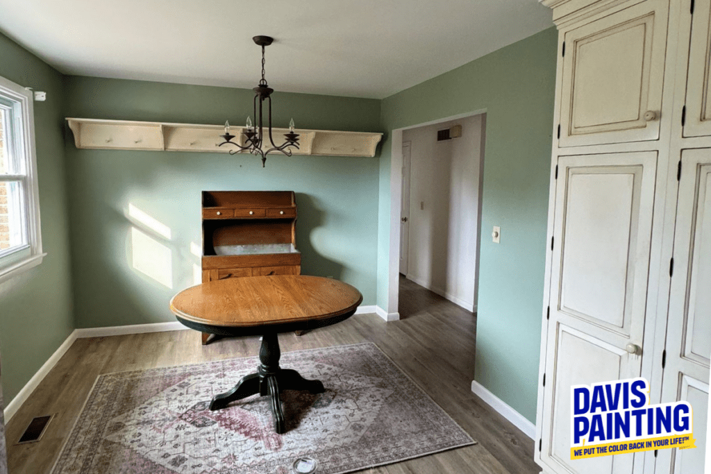 A small dining room features green walls, a round wooden table with an ornate base on a patterned rug, a wooden hutch, and a chandelier. Sunlight streams through a window. A "Davis Painting" sign is visible in the corner.