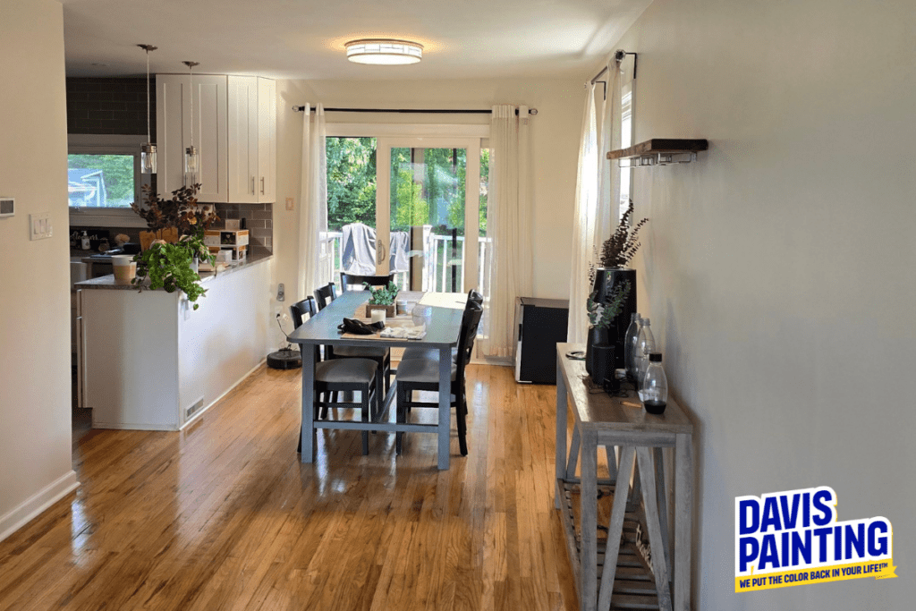 Bright dining area with a wooden table and chairs, surrounded by a white-walled interior. Natural light pours in through large patio doors, leading to a garden. A Davis Painting sign is visible in the bottom right corner.