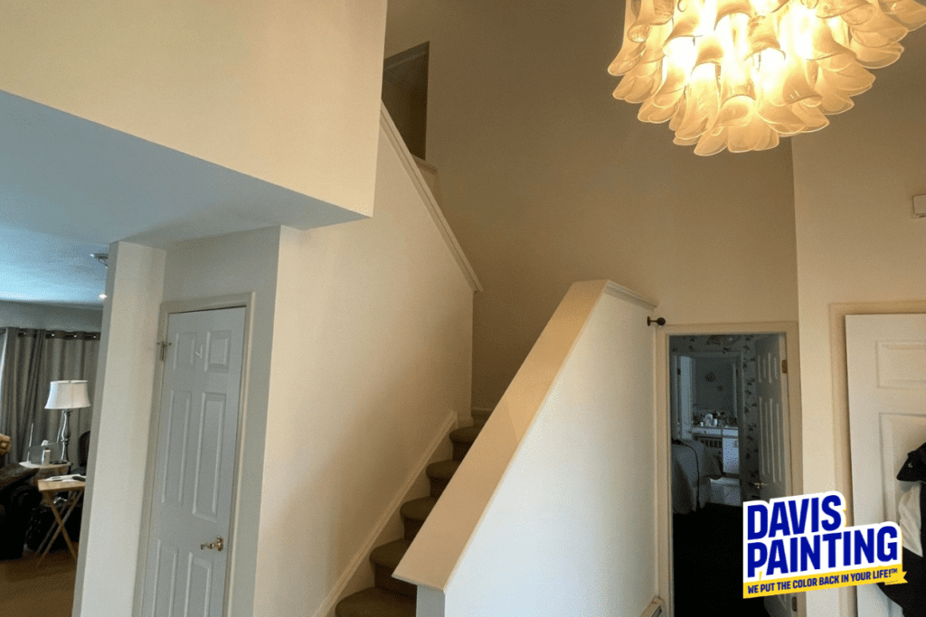 Interior view of a home featuring a staircase with beige carpet leading to an upper floor. The walls are painted white, and there's a modern chandelier above. The image includes the logo "Davis Painting" in the bottom right corner.