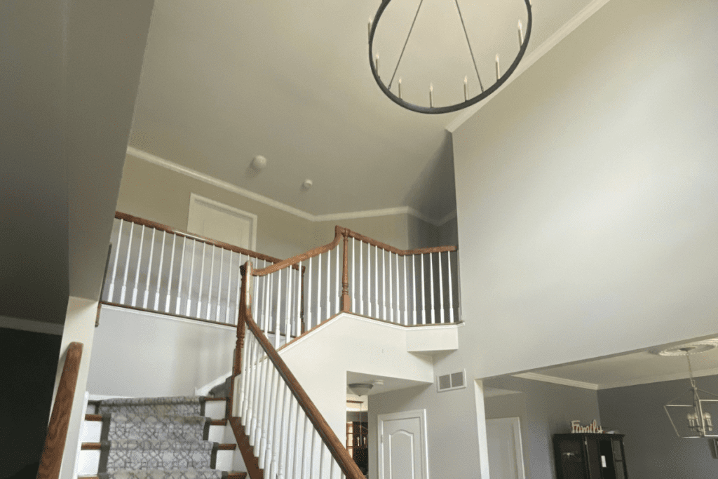 A staircase with wooden railings leads up to an upper floor with a balcony. The walls are painted light gray, and a modern chandelier hangs from the high ceiling. A decorative word "Family" can be seen on a shelf in the background.