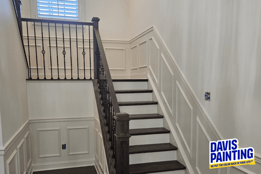 A freshly painted staircase with dark wooden steps and white wainscoting. Black metal balusters line the railing. A logo in the corner reads, "Davis Painting - We put the color back in your life!".