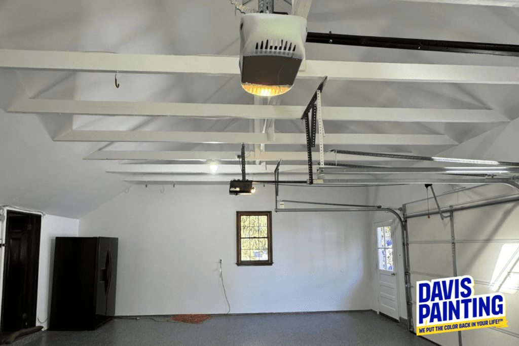 A newly painted garage interior with white walls and exposed beams. An open garage door leads outside, and a black refrigerator stands by a wooden door. The floor is gray, and a logo in the corner reads "Davis Painting.