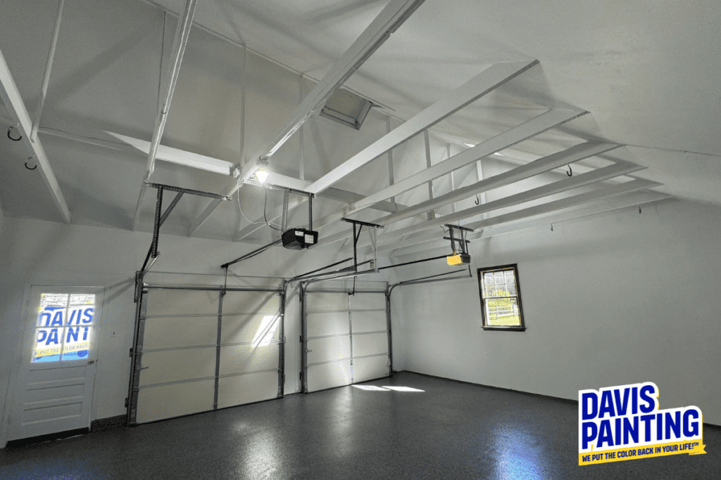 A freshly painted, spacious garage with white walls and ceiling, featuring visible rafters and a small window. Two closed garage doors dominate one wall. There's a logo for "Davis Painting" in the bottom right corner.