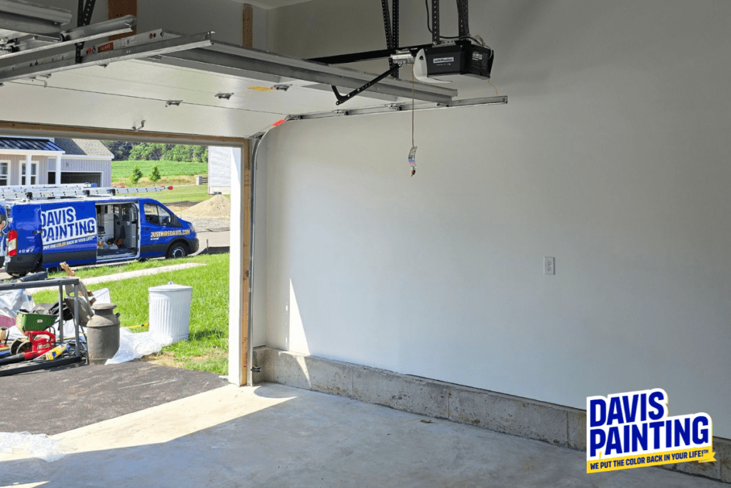 A freshly painted white garage interior with an open door. Outside, a blue Davis Painting van is parked on the driveway. The sunny weather highlights a green lawn and neighboring houses in the background.