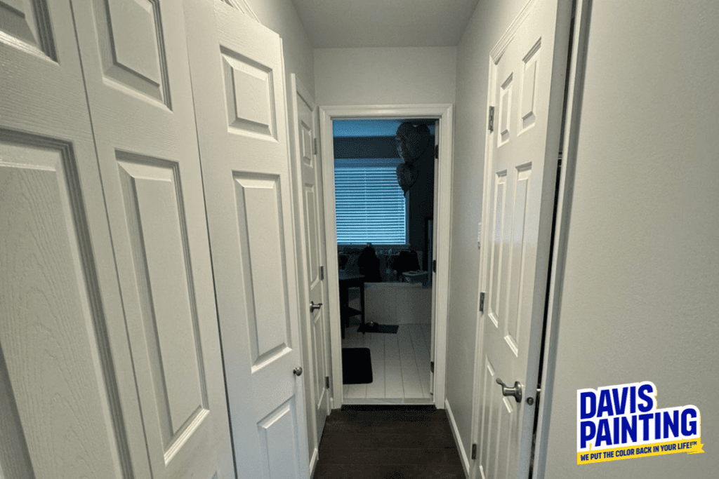 A narrow hallway with white doors on both sides, leading to a room with a window that has blinds. The walls are painted white and the floor is dark wood. A logo in the bottom right corner says "Davis Painting: We put the color back in your life!.